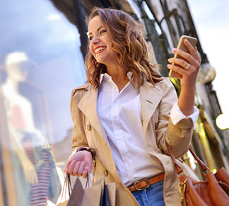 Photo of a young woman shopping in the city while checking her mobile phone and smiling.
